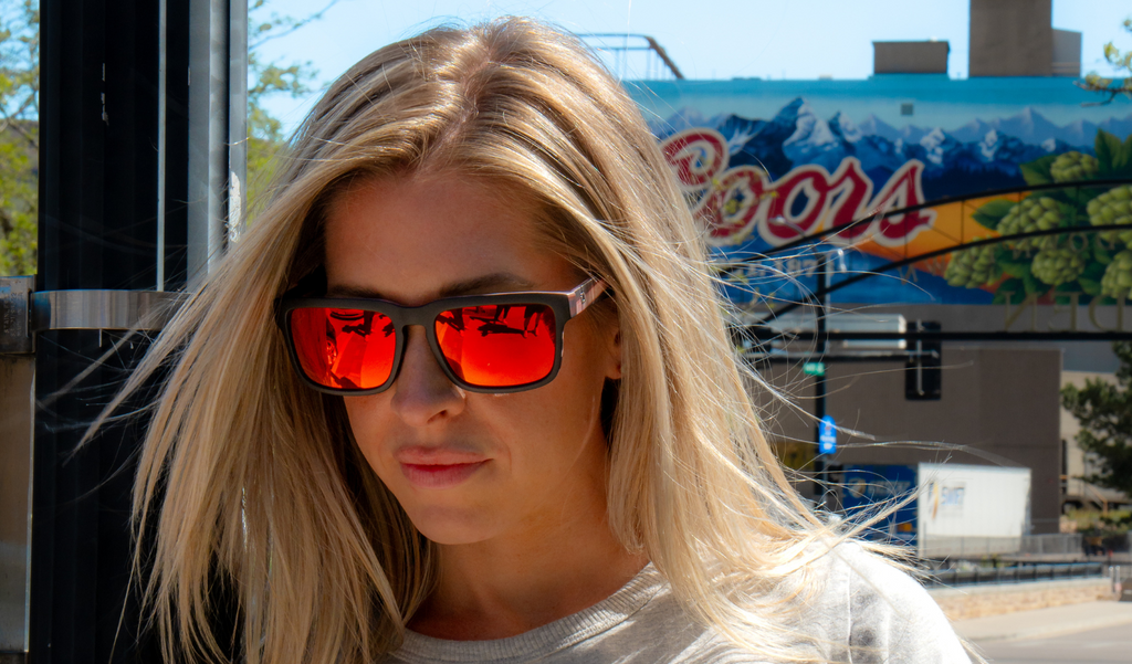 Person wearing Optic Nerve Mashup sunglasses with a black frame, smoke lenses, and a red mirror with a blurred background.