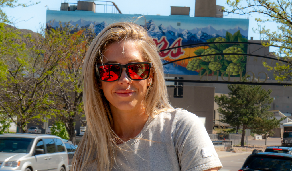 Person wearing Optic Nerve Mashup sunglasses with a black frame, smoke lenses, and a red mirror, with a colorful billboard in the background