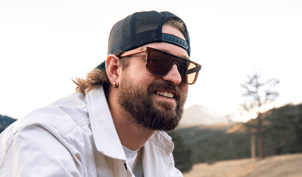 Man wearing Optic Nerve Mojo Filter Wood sunglasses with FSC wood temples, tortoise frame, and brown lens, wearing a white shirt with blurred trees and sky background