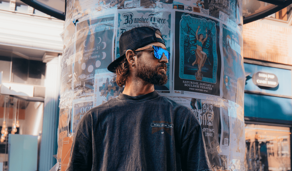 Man wearing Optic Nerve Mojo Filter Wood sunglasses with FSC wood temples, black frame, smoke lens, and blue mirror with newspaper pole background