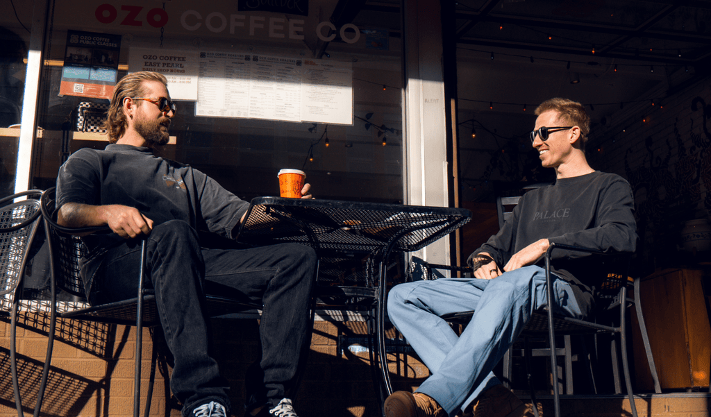 Two men sitting at an outdoor table wearing Optic Nerve sunglasses