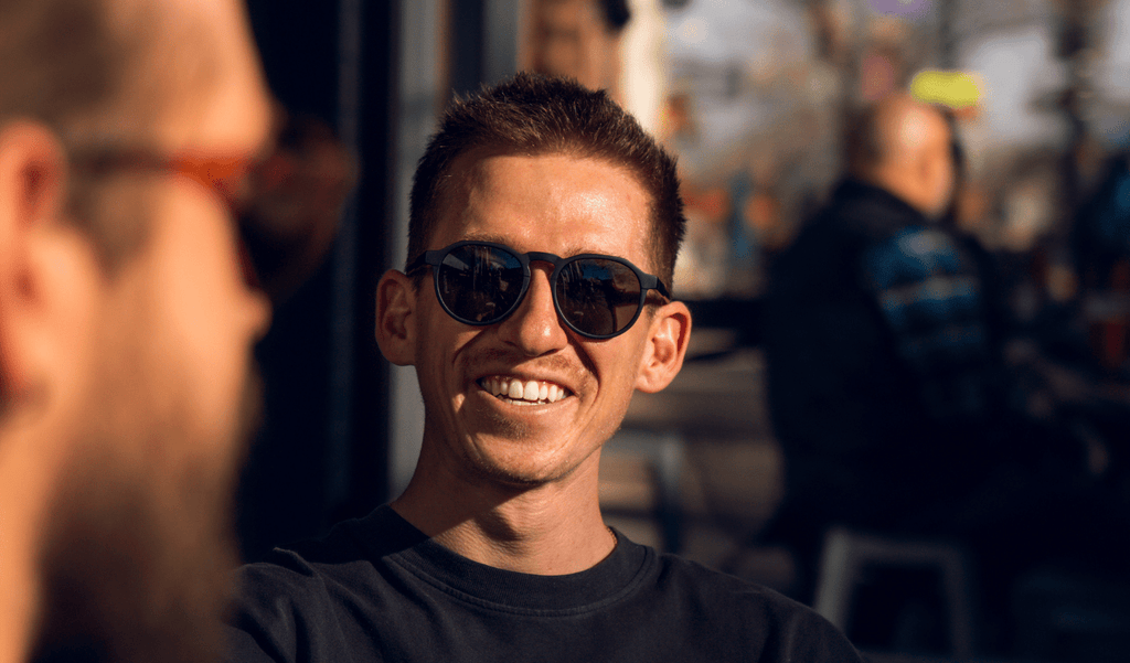 Man wearing Optic Nerve RiNO sunglasses with black frame and smoke lens, smiling with a blurred background