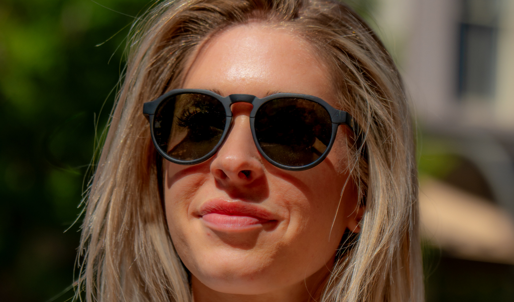 Woman wearing Optic Nerve RiNO sunglasses with a black frame and smoke lens, with a blurred outdoor background