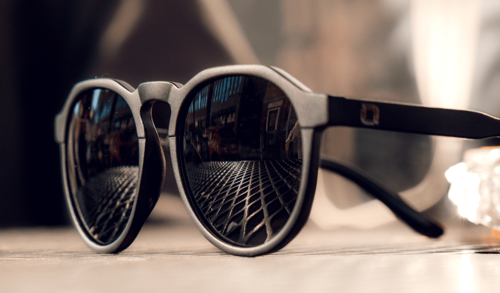 Close up of Optic Nerve RiNO sunglasses with a black frame and smoke lens resting on a grate table with a blurred background