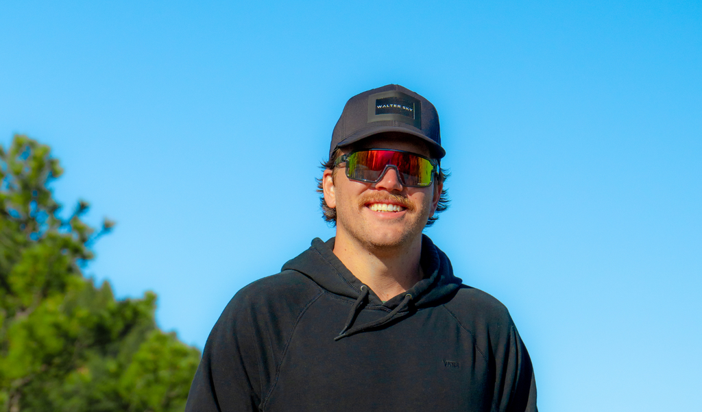 Person wearing a cap and Optic Nerve Scraggy sunglasses with a gray frame, smoke lens, and a red mirror, with a clear blue sky and trees in the background