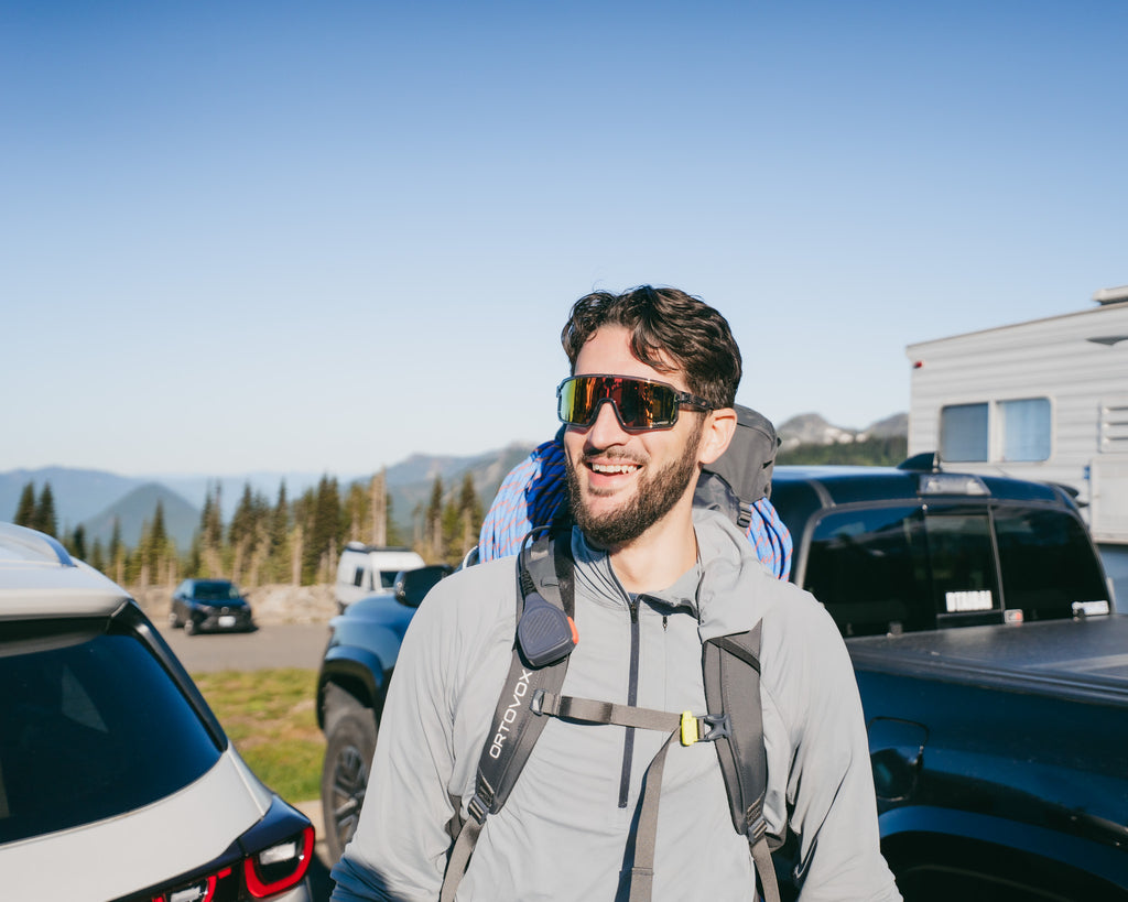 Man with Optic Nerve Scraggy sunglasses with a gray frame, smoke lens, and red mirror, and backpack standing in front of vehicles and mountains