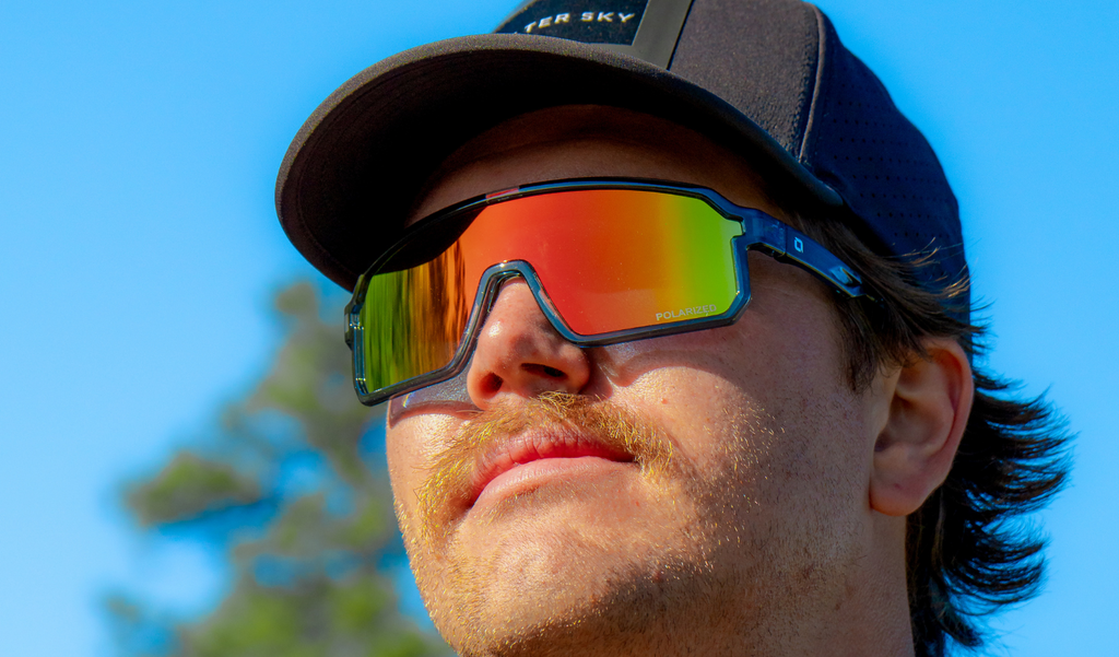 Person wearing a cap and Optic Nerve Scraggy sunglasses with a gray frame, smoke lens, and a red mirror, with a clear blue sky background
