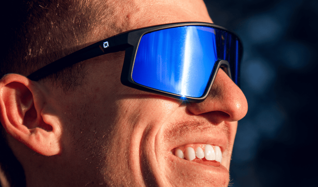 Close up of a man wearing Optic Nerve Stone Cat sunglasses with a black frame, brown lens, and a blue mirror with a blurred  background