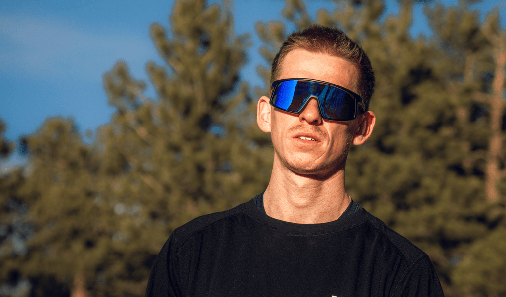 Man wearing Optic Nerve Stone Cat sunglasses with a black frame, brown lens, and a blue mirror with blurred trees in the background