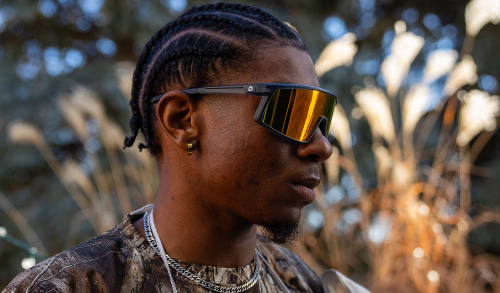 Profile of a man wearing Optic Nerve Stone Cat sunglasses with a gray frame, smoke lens, and a red mirror on a natural background