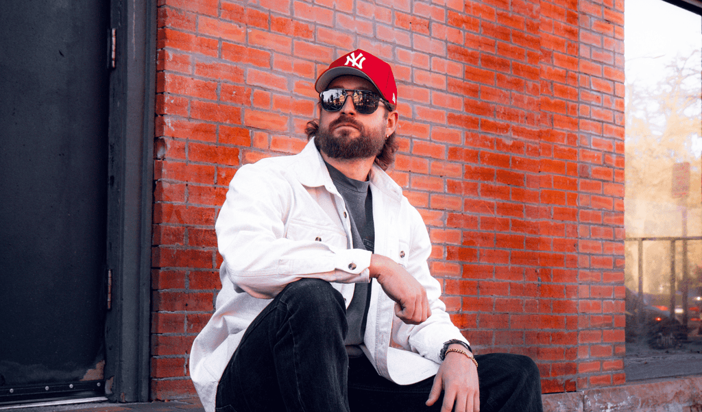 Man wearing a red New York Yankees baseball cap and Optic Nerve Wonderspoon sunglasses with a gray frame and smoke lenses in front of a  brick wall