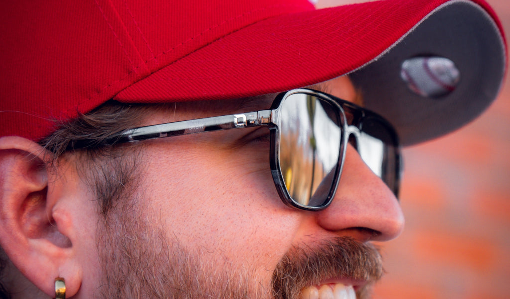 Close up of a man wearing a red baseball cap and Optic Nerve Wonderspoon sunglasses with a gray frame and smoke lenses.