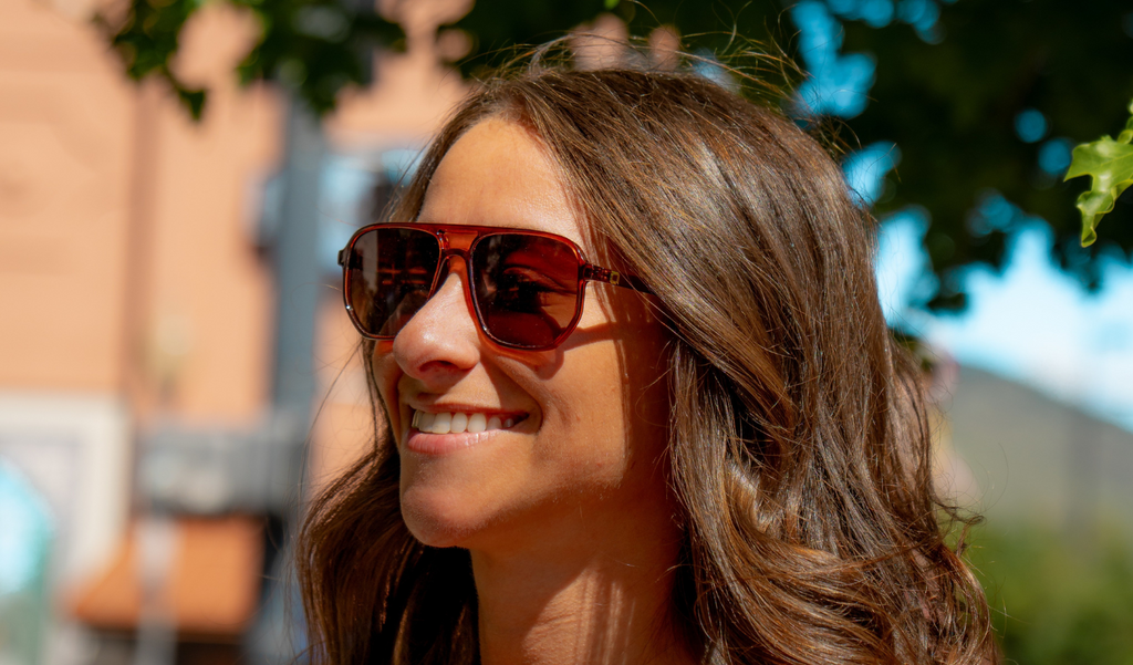 Woman wearing Optic Nerve Wonderspoon sunglasses with a brown frame and brown lenses, with a blurred outdoor background