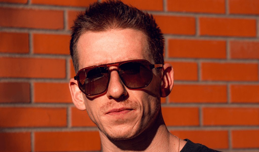 Man wearing Optic Nerve Wonderspoon sunglasses with a brown frame and brown lenses with a brick wall background.