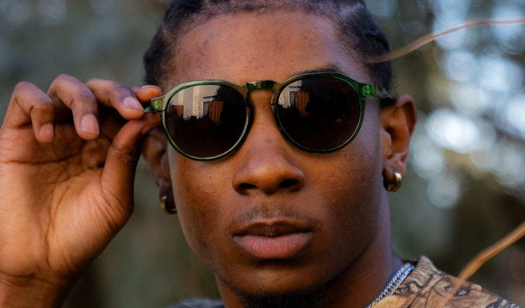 Close up of a man wearing Optic Nerve RiNO sunglasses with a green frame and brown lens with a blurred background