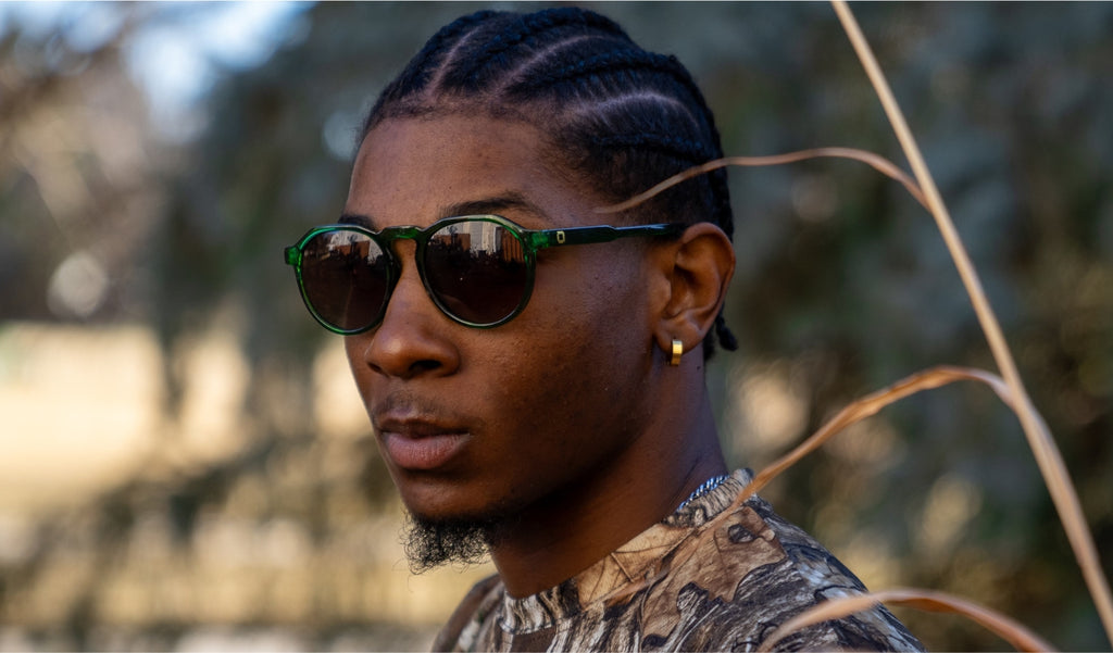 Man wearing Optic Nerve RiNO sunglasses with a green frame and brown lens with a blurred natural background