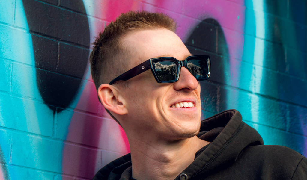 Man wearing Optic Nerve Afterglow sunglasses with a black frame and smoke lenses with a colorful graffiti wall background