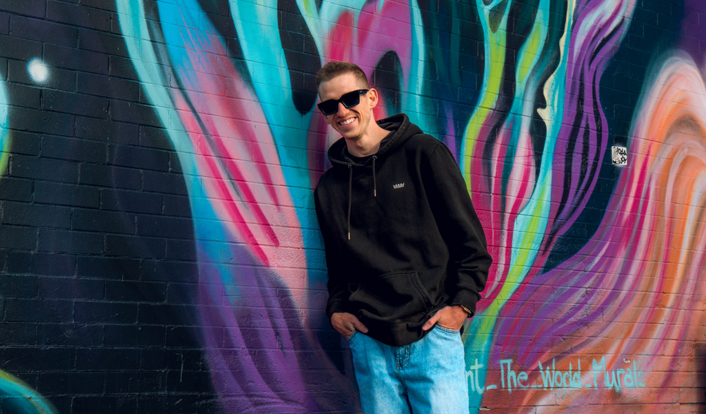 Person wearing Optic Nerve Afterglow sunglasses with a black frame and smoke lenses, and a black hoodie standing in front of a colorful mural.