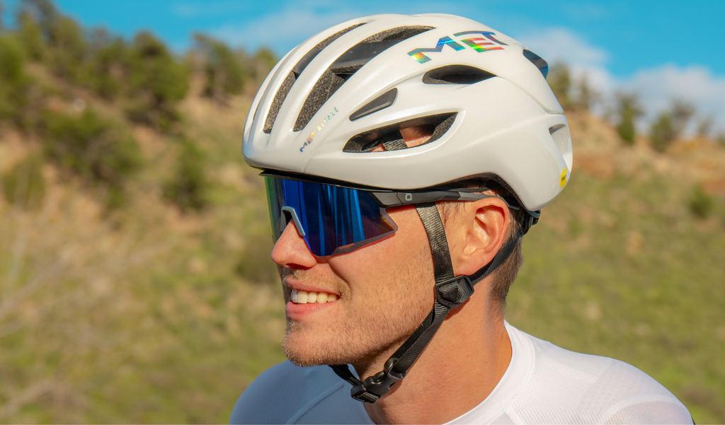 Person wearing a white helmet with a colorful logo and Optic Nerve FixieAIR sunglasses with a black frame, brown lens, and blue mirror, outdoors