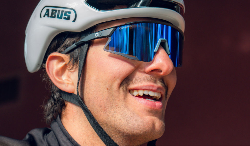 Person wearing a white helmet with 'ABUS' branding and Optic Nerve FixieAIR Sunglasses with a black frame, brown lens, and a blue mirror, against a blurred background.