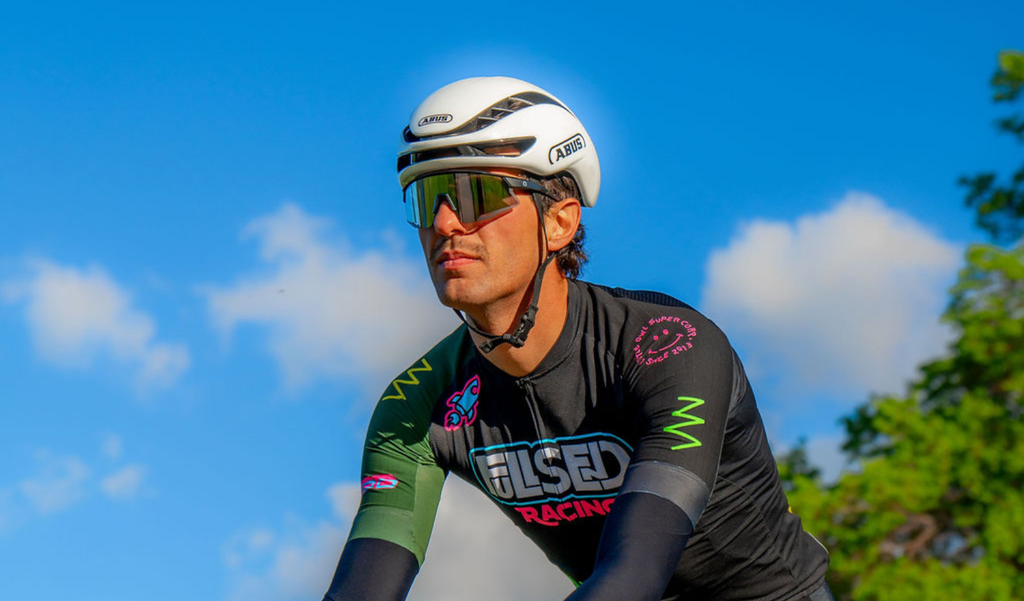 Cyclist wearing a helmet and Optic Nerve FixieAIR sunglasses with a black frame, smoke lens, and a silver mirror, with a blue sky and trees in the background