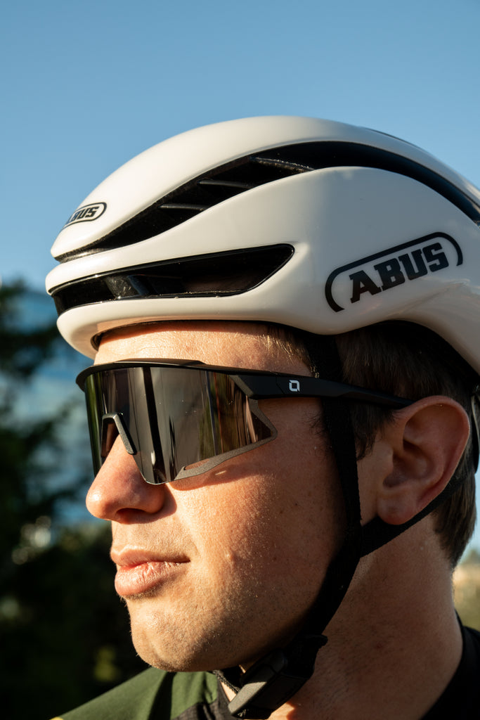 Cyclist wearing a white helmet and FixieAIR sunglasses with a black frame, smoke lens, and silver mirror on a blurred background