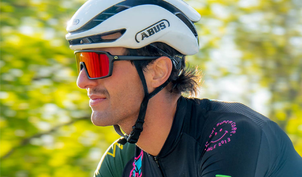 Cyclist wearing a white helmet and Optic Nerve FixiePHENOM 2.0 Photochromic sunglasses with a black frame, photochromic lens, and red mirror on a blurred background