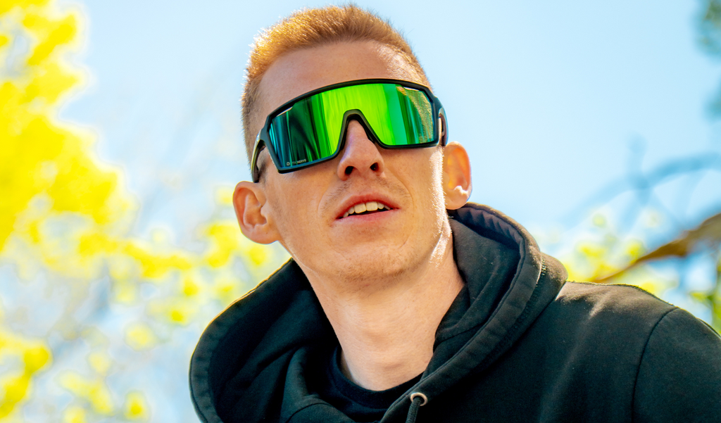 Man wearing Optic Nerve FixiePHENOM 2.0 sunglasses with a black frame, smoke lens, and a green mirror with a blurred background