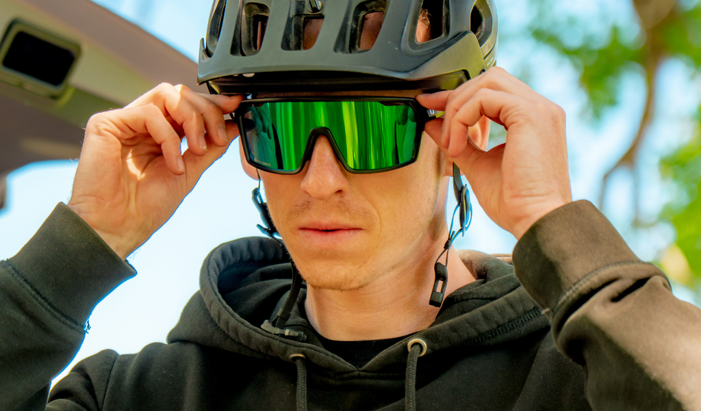Cyclist wearing a black helmet and FixiePHENOM 2.0 sunglasses with a black frame, smoke lens, and a green mirror with a blurred background
