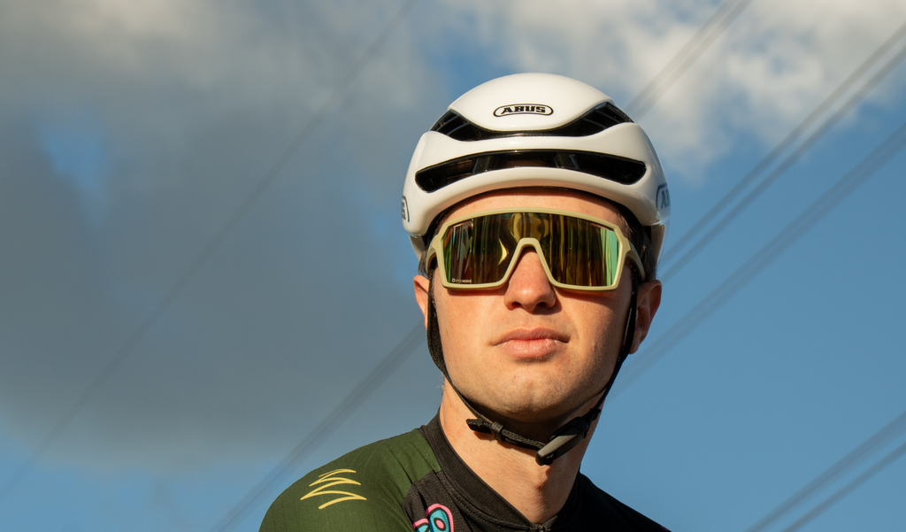 Cyclist wearing a white helmet a Optic Nerve FixiePHENOM 2.0 sunglasses with a green frame, brown lens, and a gold mirror with a blue sky background