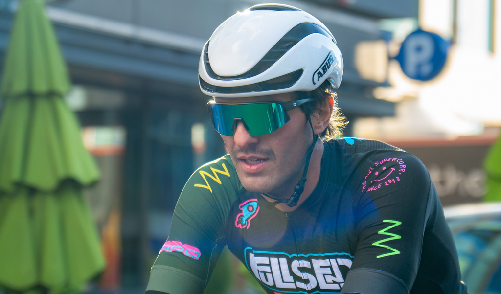 Cyclist wearing a white helmet and Optic Nerve FixieSLICE sunglasses with a gray frame, smoke lens, and a green mirror with a blurred downtown background