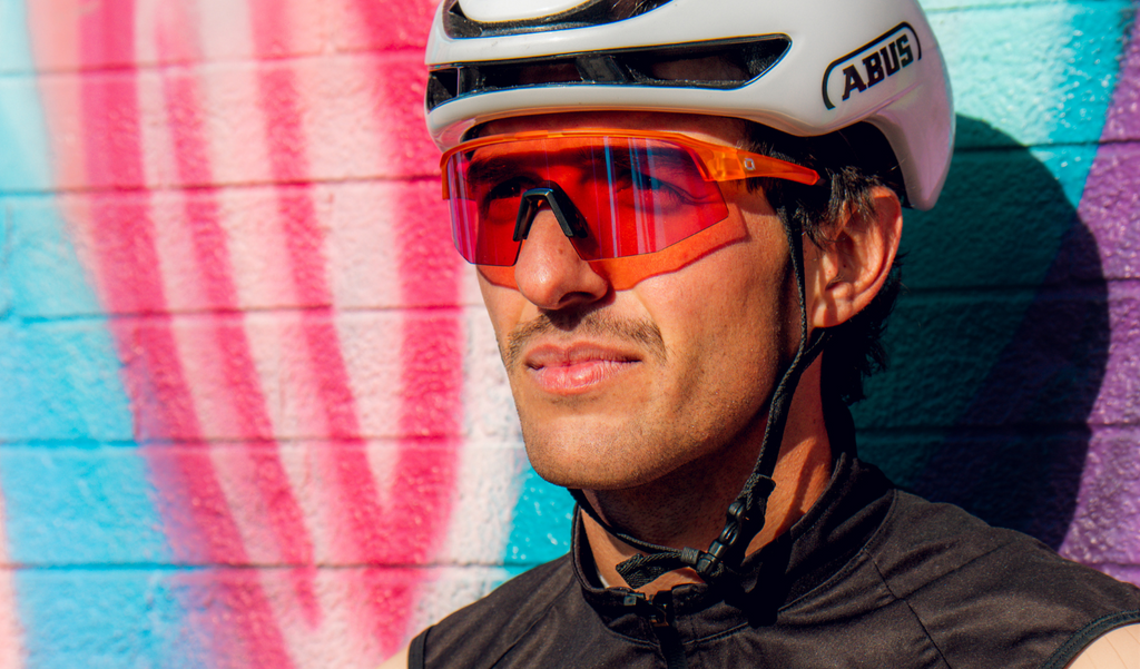 Cyclist wearing a white helmet and FixieSLICE sunglasses with an orange frame, rose lens, and a blue mirror with a mural background