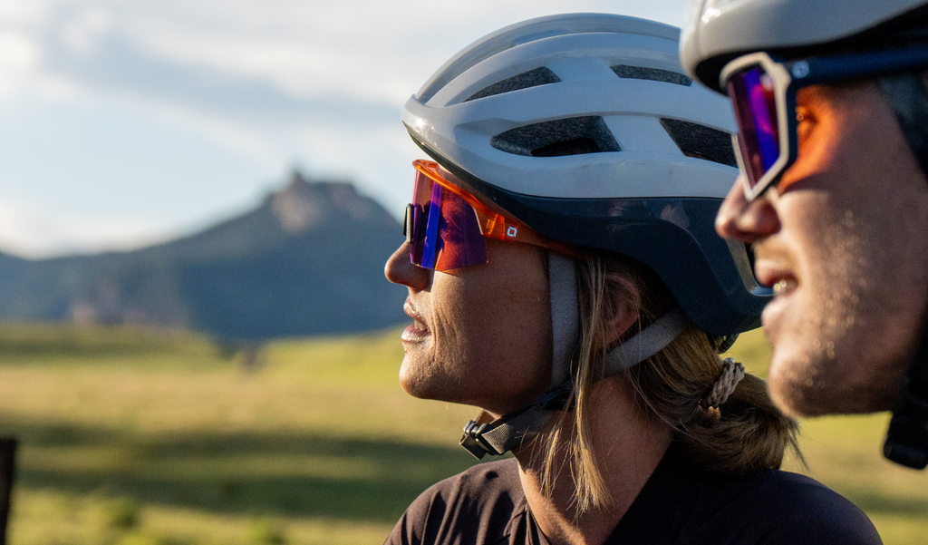 Cyclist wearing a white helmet and Optic Nerve FixieSLICE sunglasses with an orange frame, rose lens, and a blue mirror, with a mountain in the background