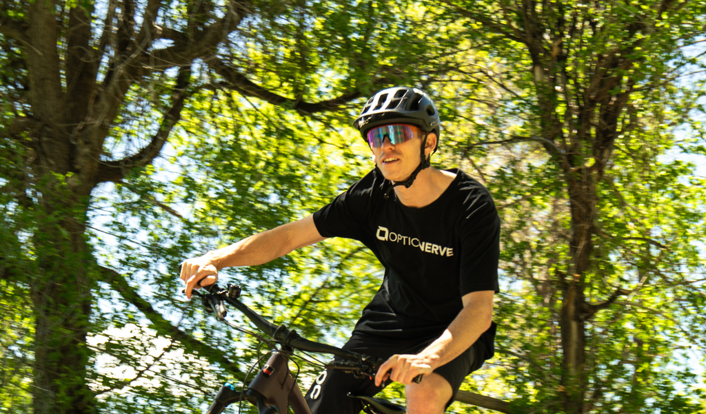 Cyclist wearing a black helmet, FixieSLICE sunglasses with an orange frame, rose lens, and a blue mirror, with an Optic Nerve shirt riding a black bike with a blurred tree background