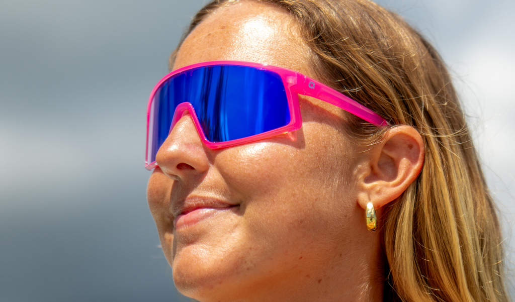 Person wearing Optic Nerve Gravel Cat sunglasses with a pink frame, brown lens, and a blue mirror, with a blurred background