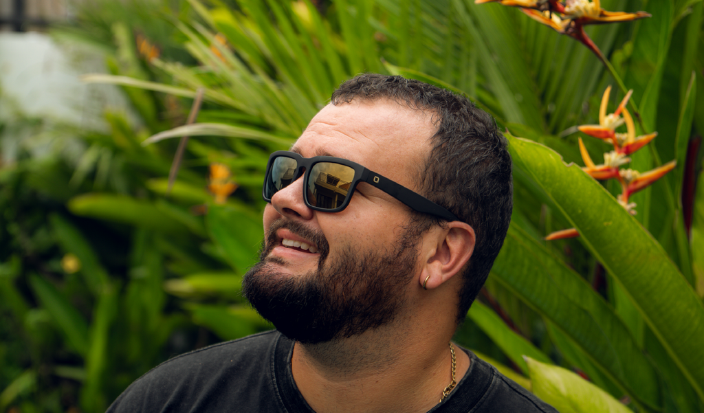 Man wearing Optic Nerve Mashup sunglasses with a black frame, brown lenses, and a gold mirror with a tropical plant background