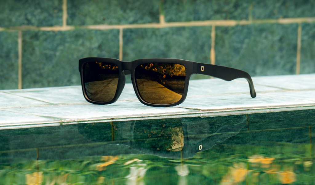 Optic Nerve Mashup sunglasses with a black frame, brown lenses, and a gold mirror on a pool ledge.