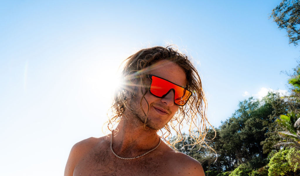 Man wearing Optic Nerve Mojo Filter sunglasses with a black frame, smoke lens, and red mirror with a tropical background and the sun shinning