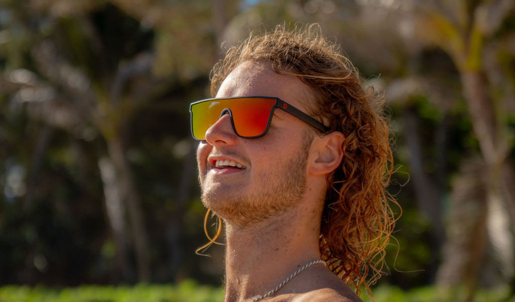 Man wearing Optic Nerve Mojo Filter sunglasses with a black frame, smoke lens, and red mirror with a blurred tropical background