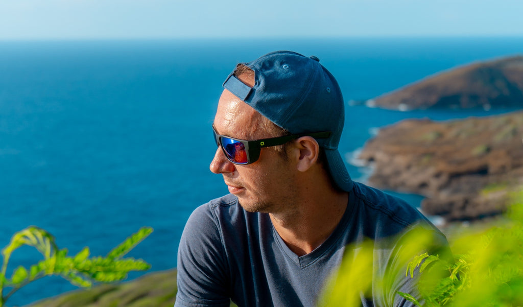 Man wearing a blue baseball cap and Optic Nerve Staunton sunglasses with a black frame, brown lenses, ad a blue mirror with a blurred ocean background.