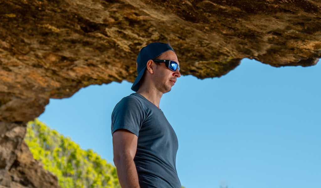 Man wearing a blue baseball cap and Optic Nerve Staunton sunglasses with a black frame, brown lenses, and a blue mirror with a blurred rocky background.