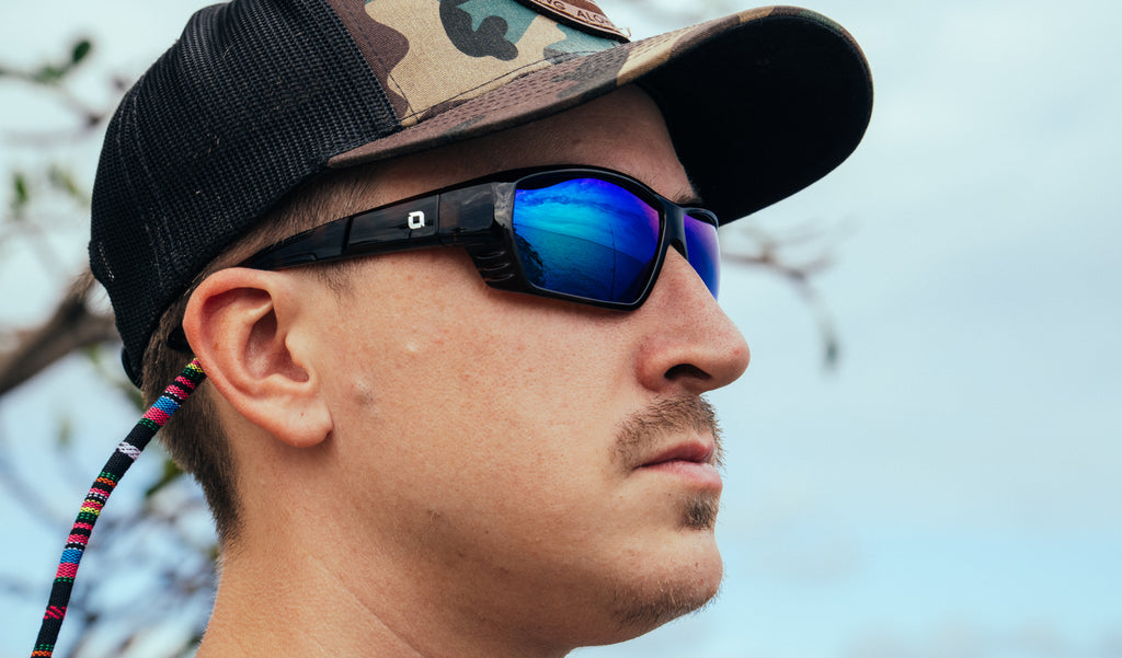Close up of a man wearing a cap and Optic Nerve Yampa sunglasses with a tortoise frame, brown lenses, and a green mirror with a blurred background.
