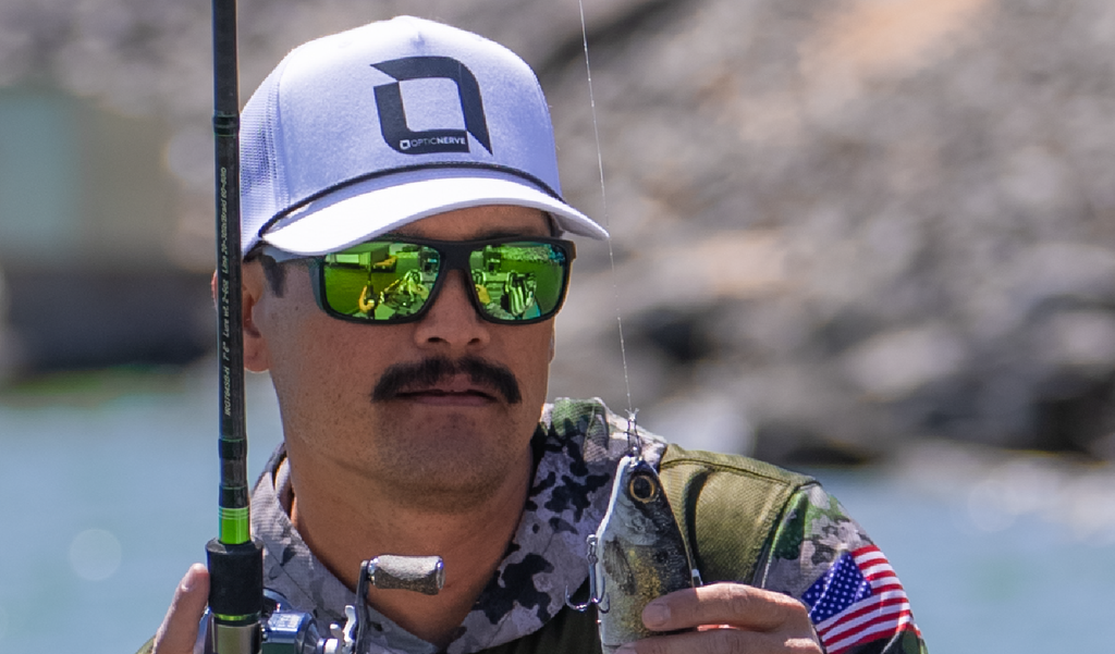 Man wearing an Optic Nerve cap and Optic Nerve Zaldaingerous Cuda sunglasses with a black frame, amber lenses, and green mirror, holding a fishing rod with a blurred natural background