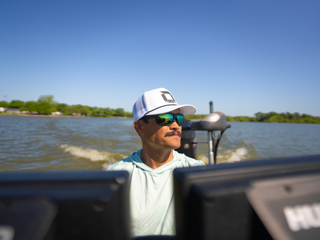 Man on a boat wearing Zaldaingerous Cuda sunglasses and an Optic Nerve cap, with water and trees in the background