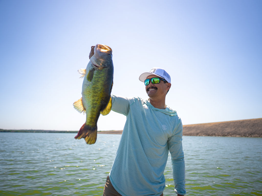 A man holding a large fish by a lake wearing the Zaldaingerous Cuda Sunglasses with clear blue sky