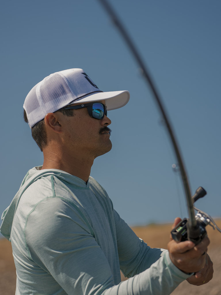 A fisherman in a white Optic Nerve baseball cap wearing the Zaldaingerous Cuda Sunglasses casting a fishing line on a blurred background