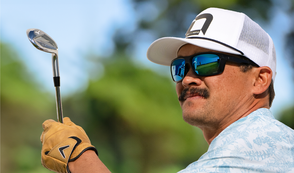 A golfer wearing an Optic Nerve baseball cap and a pair of Zaldaingerous Cuda Sunglasses with a black frame, amber lens, and blue mirror holding a golf club on a blurred background