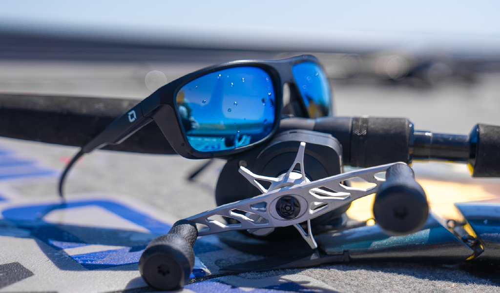 Fishing rod with Optic Nerve Zaldaingerous Cuda Sunglasses; black frame, amber lenses, and a blue mirror, on a blurred background