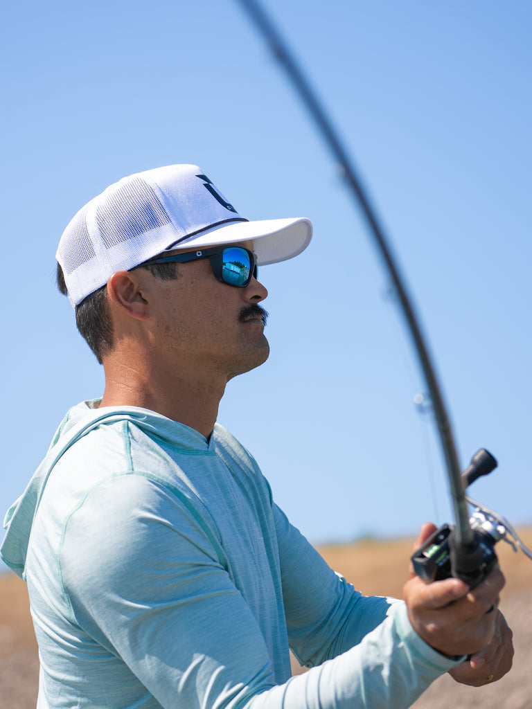 A fisherman in a white Optic Nerve baseball cap wearing the Zaldaingerous Cuda Sunglasses casting a fishing line on a blurred background