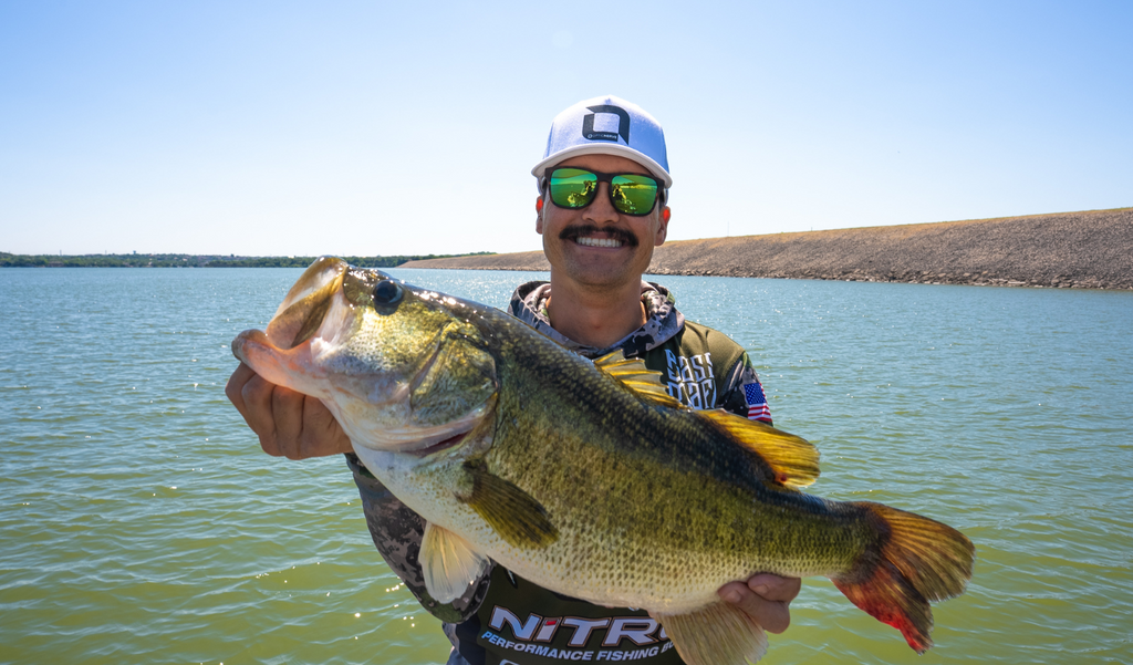 Chris Zaldain wearing an Optic Nerve cap, Optic Nerve Zaldaingerous Rumble sunglasses with a tortoise frame, amber lens, and a green mirror holding a fish with a lake background